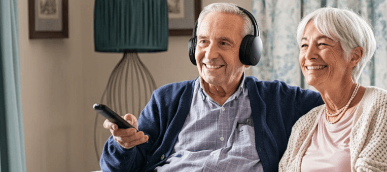 Smiling senior couple wearing wireless TV headphones while watching television together, enjoying clear sound without increasing the volume