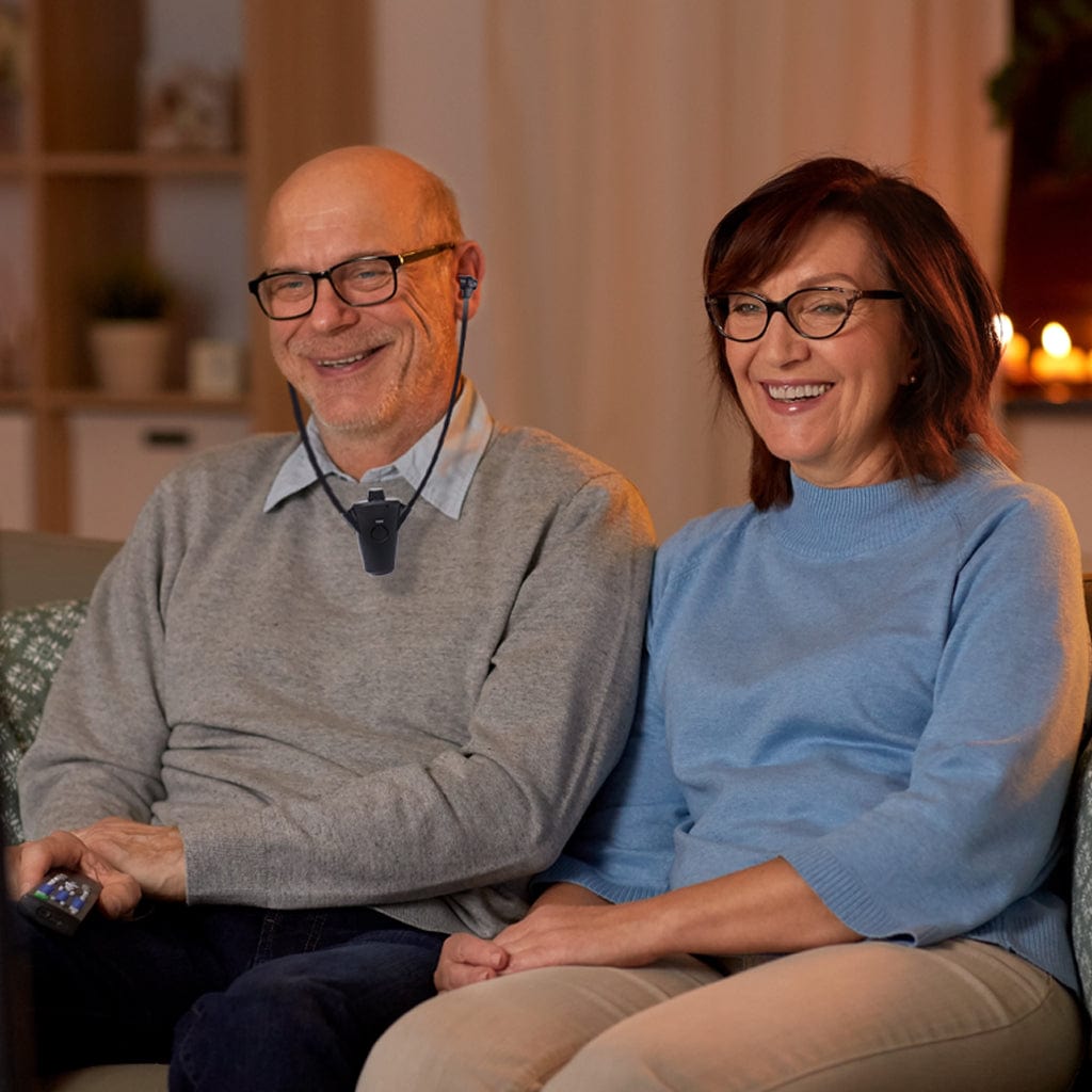 Smiling senior couple watching television while using a wireless TV listening device, enjoying clear audio and shared entertainment at home