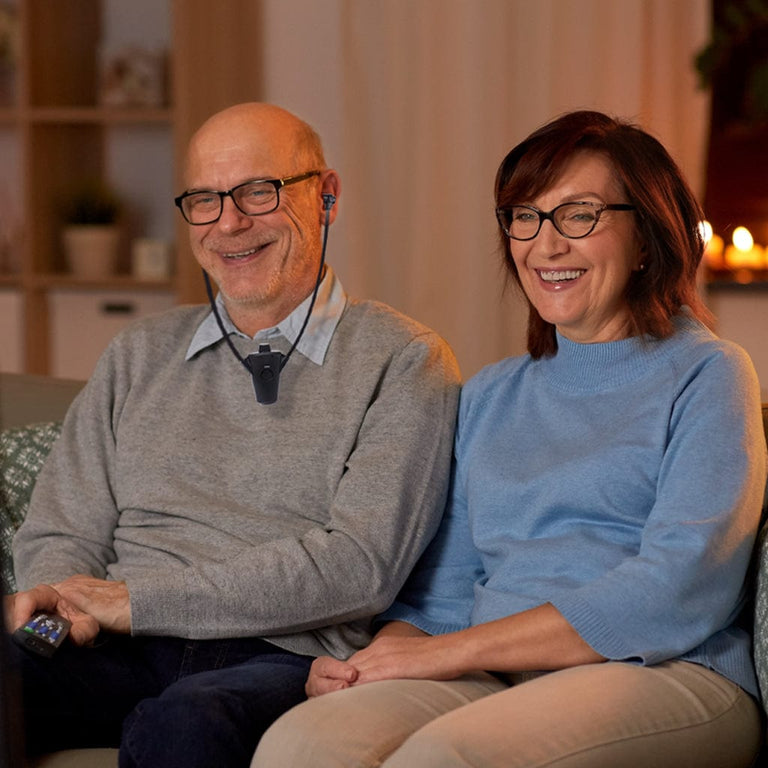 Smiling senior couple watching television while using a wireless TV listening device, enjoying clear audio and shared entertainment at home